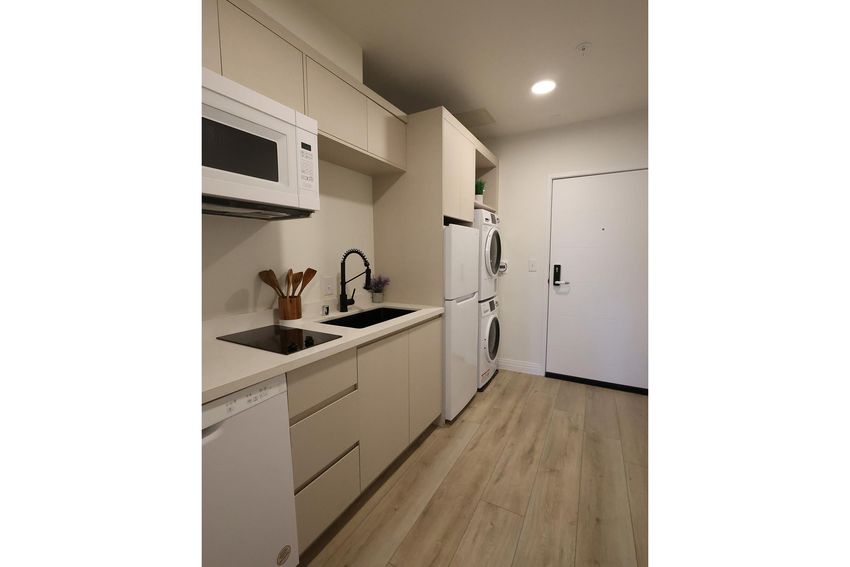 A modern kitchen featuring beige cabinetry, a microwave, a stovetop, and a dishwasher. Adjacent to the kitchen area are stacked washer and dryer units. The floor is light wood, and there's a door leading outside, adding to the functional and stylish design of the space.