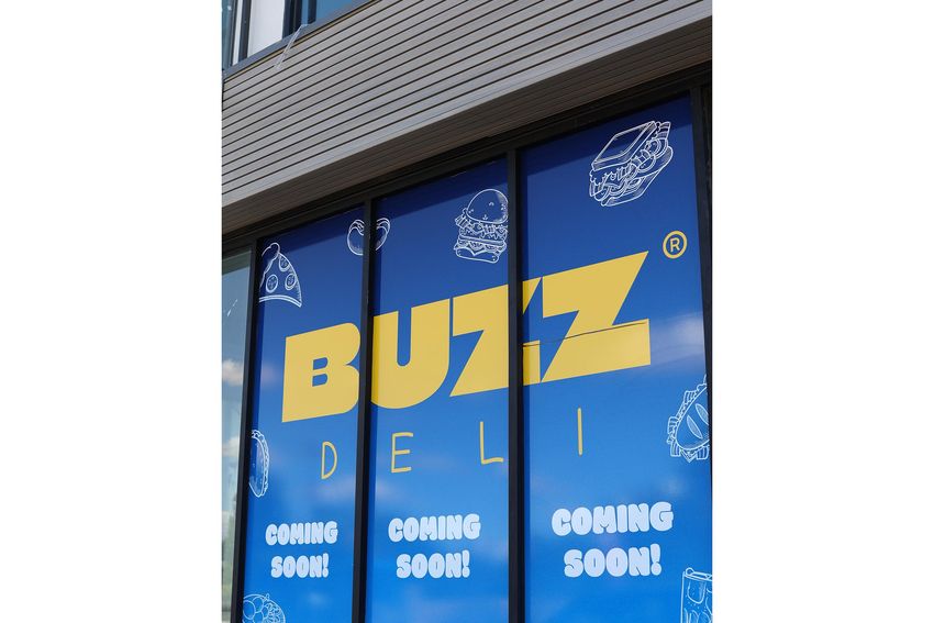 Signage for "BUZZ DELI" with a bright blue background featuring the text "COMING SOON!" The design includes illustrated food items like burgers and pizza, emphasizing the deli's offerings. This window display indicates that the establishment is preparing to open soon.