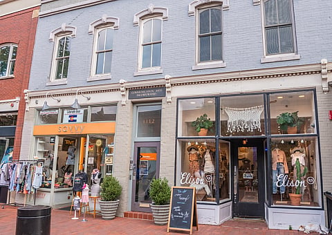 Charming street view of a brick-lined boutique with stylish storefronts, showcasing elegant clothing displays and vibrant potted plants.