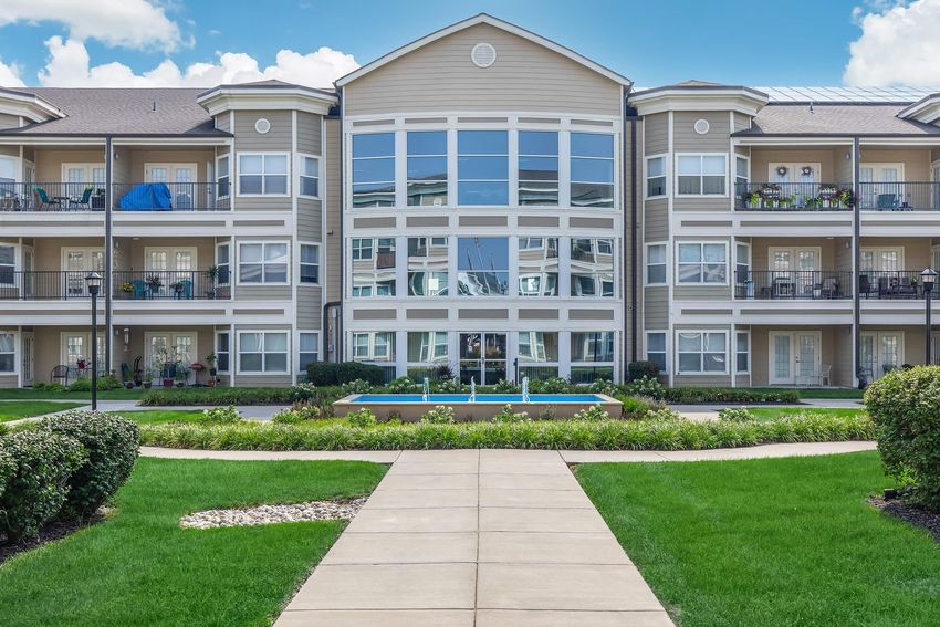 A modern multi-story residential complex with large windows and balconies, surrounded by manicured green lawns and walking paths. The central area features a small water fountain. Bright blue sky with scattered clouds adds to the cheerful atmosphere.