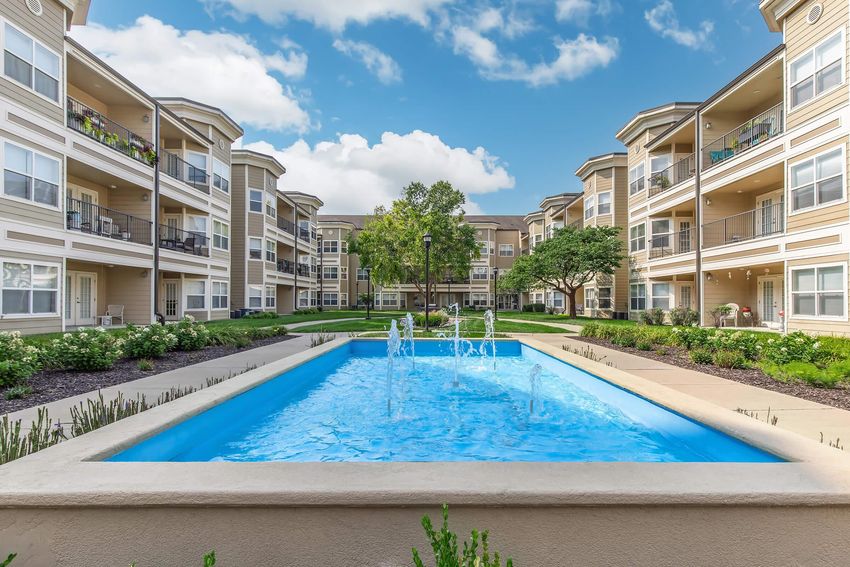 A picturesque courtyard featuring a blue fountain surrounded by landscaped greenery and multiple apartment buildings. The sky is partly cloudy, adding a bright atmosphere to the serene setting.