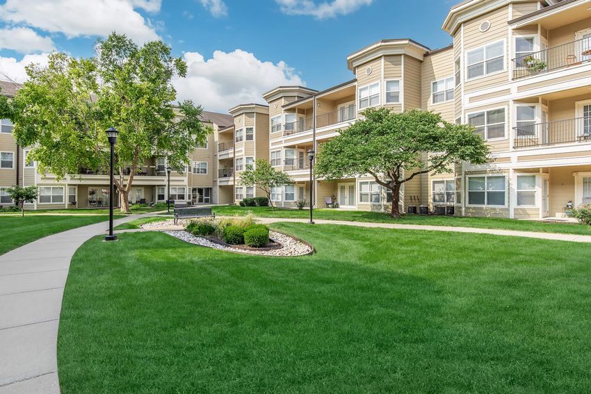 A well-maintained apartment community featuring multiple buildings with balconies. The landscape is lush with green grass, decorative shrubs, and a winding pathway. The sky is partly cloudy, allowing sunlight to illuminate the area, creating a pleasant residential environment.