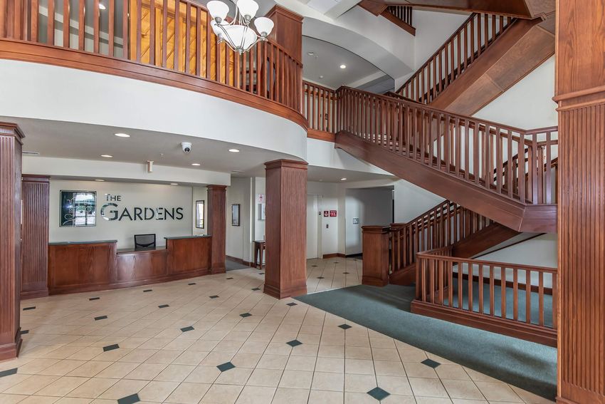 Interior view of a welcoming reception area featuring wooden stairs, a reception desk labeled "The Gardens," and decorative lighting. The space has a modern design with tiled flooring and wooden railings, creating an inviting atmosphere.