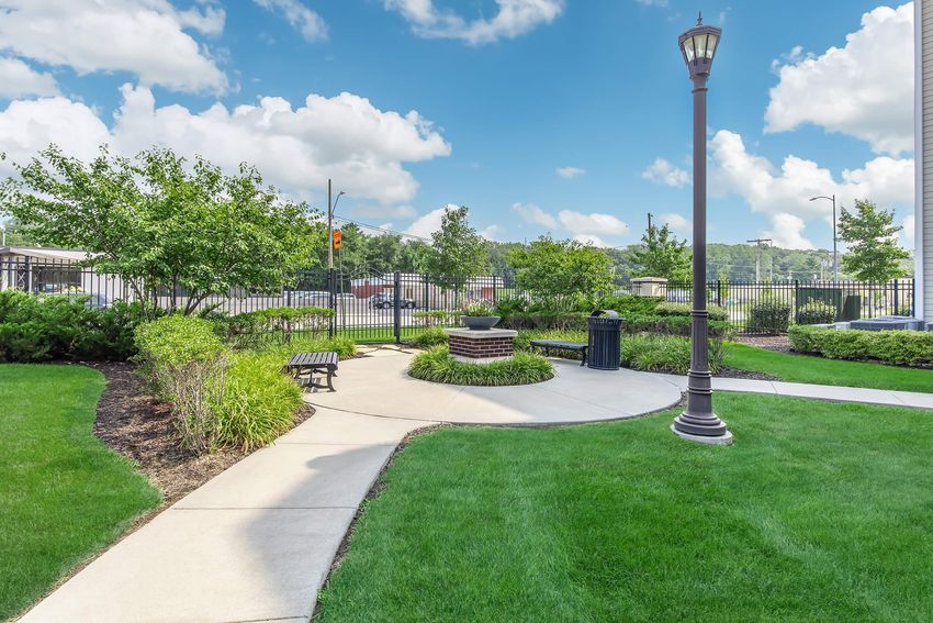 A landscaped park area featuring well-manicured grass, bushes, and trees. A winding concrete path leads to a circular stone seating area with a fire pit. A lamp post stands nearby, and in the background, a fence and some buildings can be seen under a blue sky with fluffy white clouds.