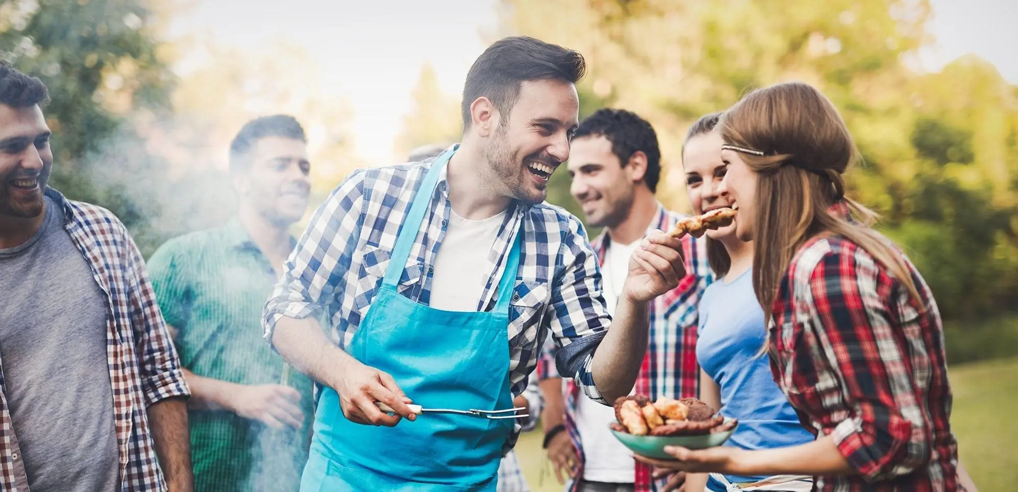 A group of people enjoying a barbecue outdoors, with one person wearing an apron and holding tongs. Others are chatting, holding food, and occasionally sharing laughs over frequently asked questions about grilling techniques.