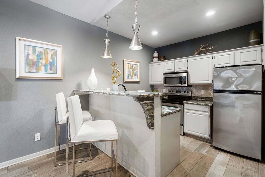 Modern kitchen featuring a gray wall, light-colored cabinetry, and stainless steel appliances. A bar area with two high-backed white stools and decorative art pieces on the walls. Stylish pendant lights hang above the bar, adding to the contemporary aesthetic. Bright and inviting atmosphere.