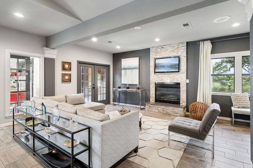 A modern living room featuring a light gray sofa, a glass coffee table, and a stone fireplace. Large windows provide natural light, while decorative elements like artwork and a cozy pouf add style. The space has a neutral color palette and a contemporary design, creating an inviting atmosphere.