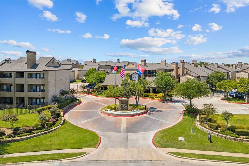Aerial view of an apartment complex with well-maintained landscaping, featuring a circular entrance and flags of the United States and Texas. The scene is bright with a blue sky and scattered clouds. Surrounding trees and flowers enhance the inviting atmosphere of the community.