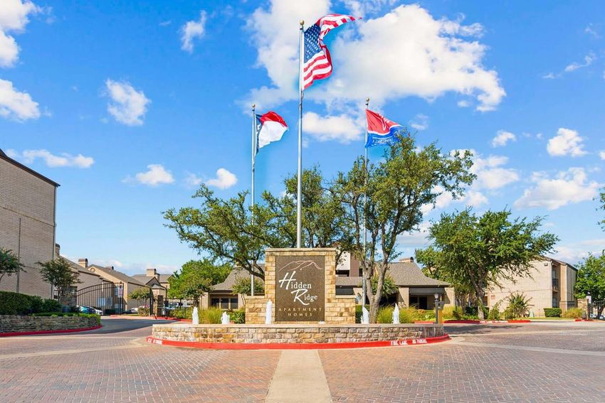 A stone entrance sign for Hidden Ridge Apartment Homes, surrounded by lush greenery and trees. Three flags are displayed in front of the sign: the American flag, a state flag, and a city flag. The sky is blue with white clouds, and the pavement is lined with red curbs.