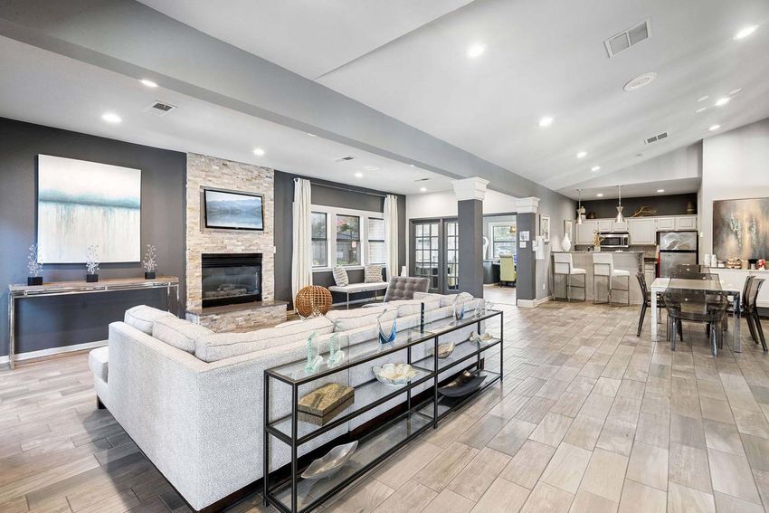 A spacious, modern living room featuring a gray sectional sofa, a stone fireplace, and large windows. The room has hardwood flooring, decorative artwork on the walls, and a stylish coffee table. A dining area is visible in the background, along with a sleek kitchen. Natural light fills the space, creating a warm atmosphere.
