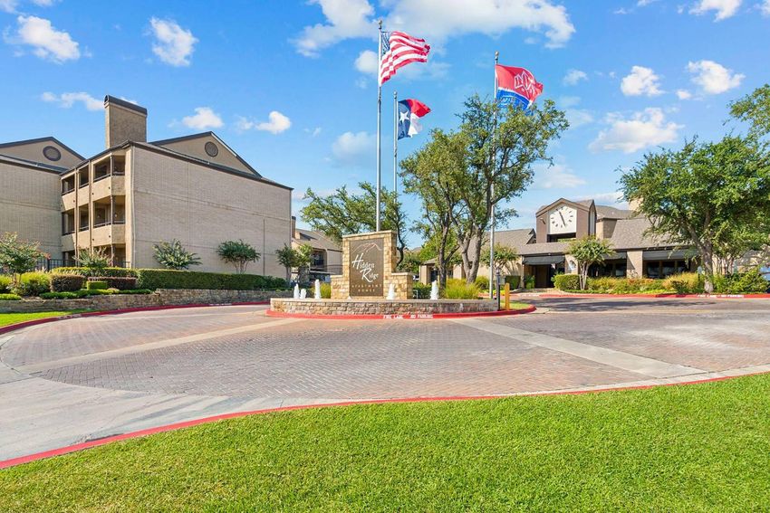Entrance to a residential community featuring a landscaped area with a stone sign, American and state flags, and modern buildings in the background. Green grass and trees are visible under a clear blue sky with scattered clouds.