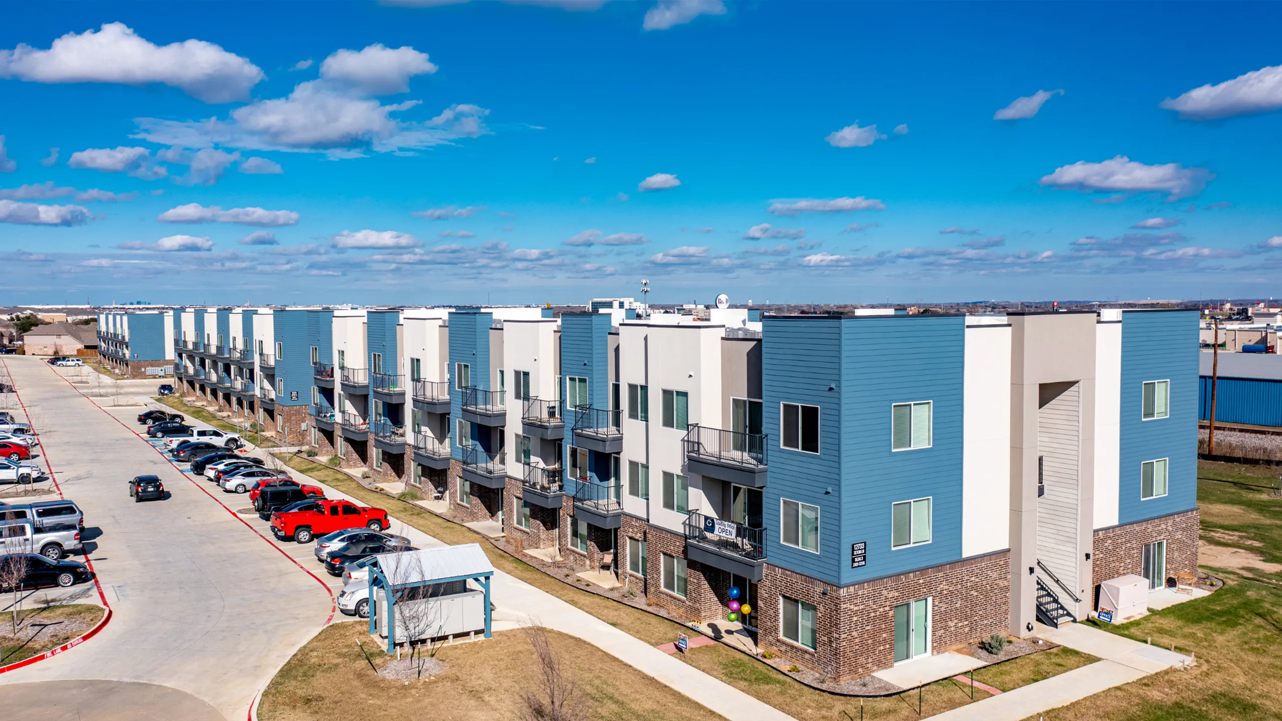 Modern apartment complex with sleek blue and white exterior, ample parking, and beautiful blue sky backdrop, perfect for convenient urban living.