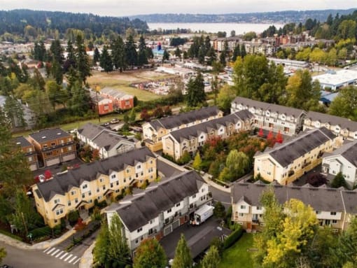 Apartments for Rent in Kenmore, WA - Northshore Townhomes - Aerial View of the Property Featuring Tan and Yellow Buildings with Grey Roofing, Surrounded by Lush Nature and Landscaping, with Nearby Lake in the Background.