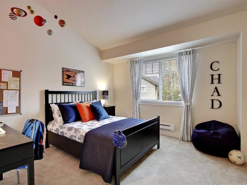 Three-Bedroom Apartments in Kenmore, WA - Northshore Townhomes - Bedroom with Carpeted Flooring, Bed, Nightstand with Lamp, Desk Chair, Bean Bag, Vaulted Ceiling, and Large Window.