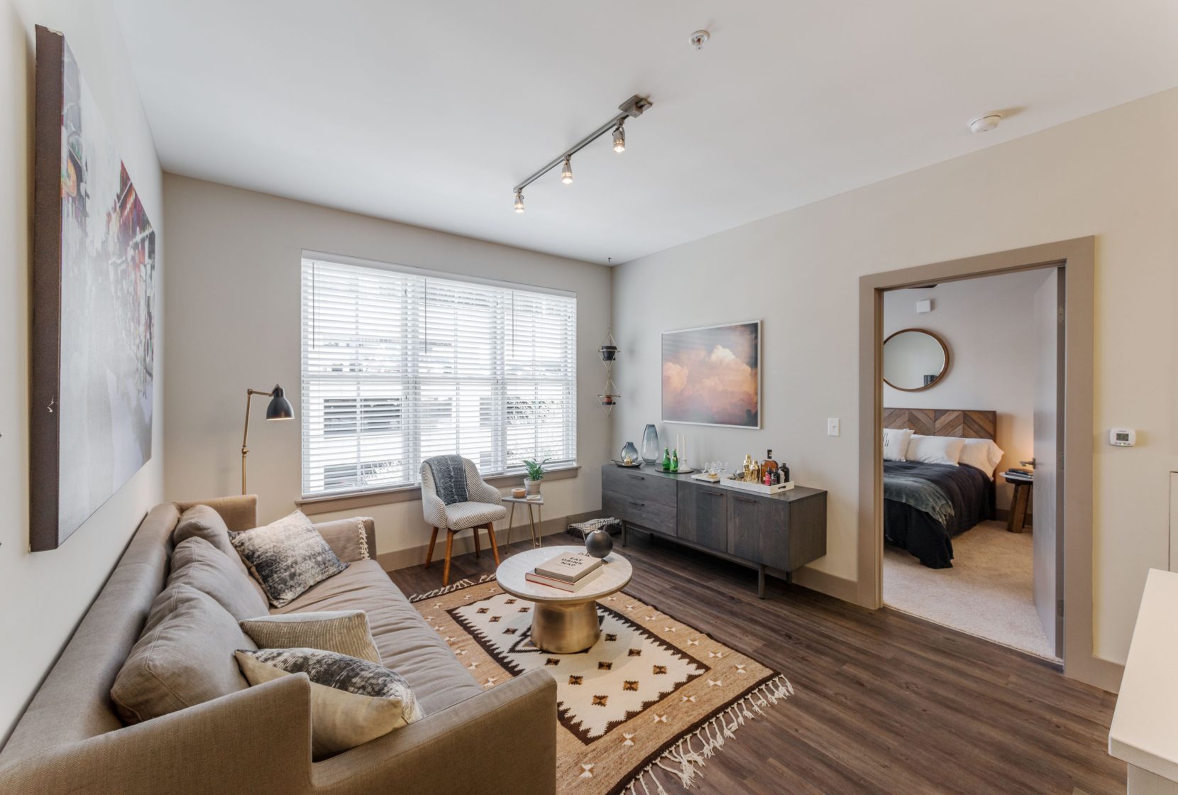 Apartment living room with large window, plank wood floors, neutral decor and walls, track lighting, access to bedroom