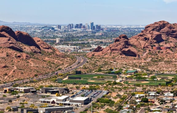 A panoramic view of the Phoenix, AZ skyline, nestled between rocky hills, with a highway winding through the landscape.