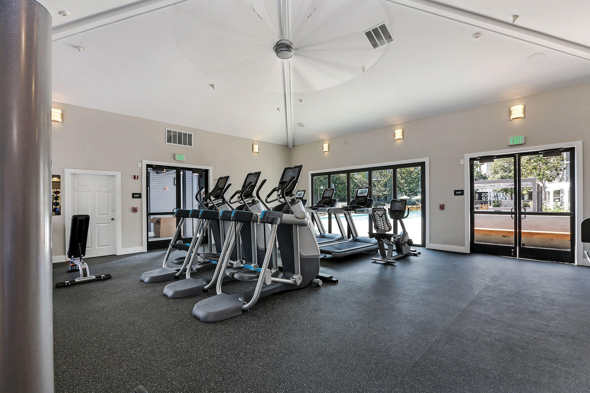 Indoor fitness center with cardio equipment facing large glass windows and doors with a view to the pool outside
