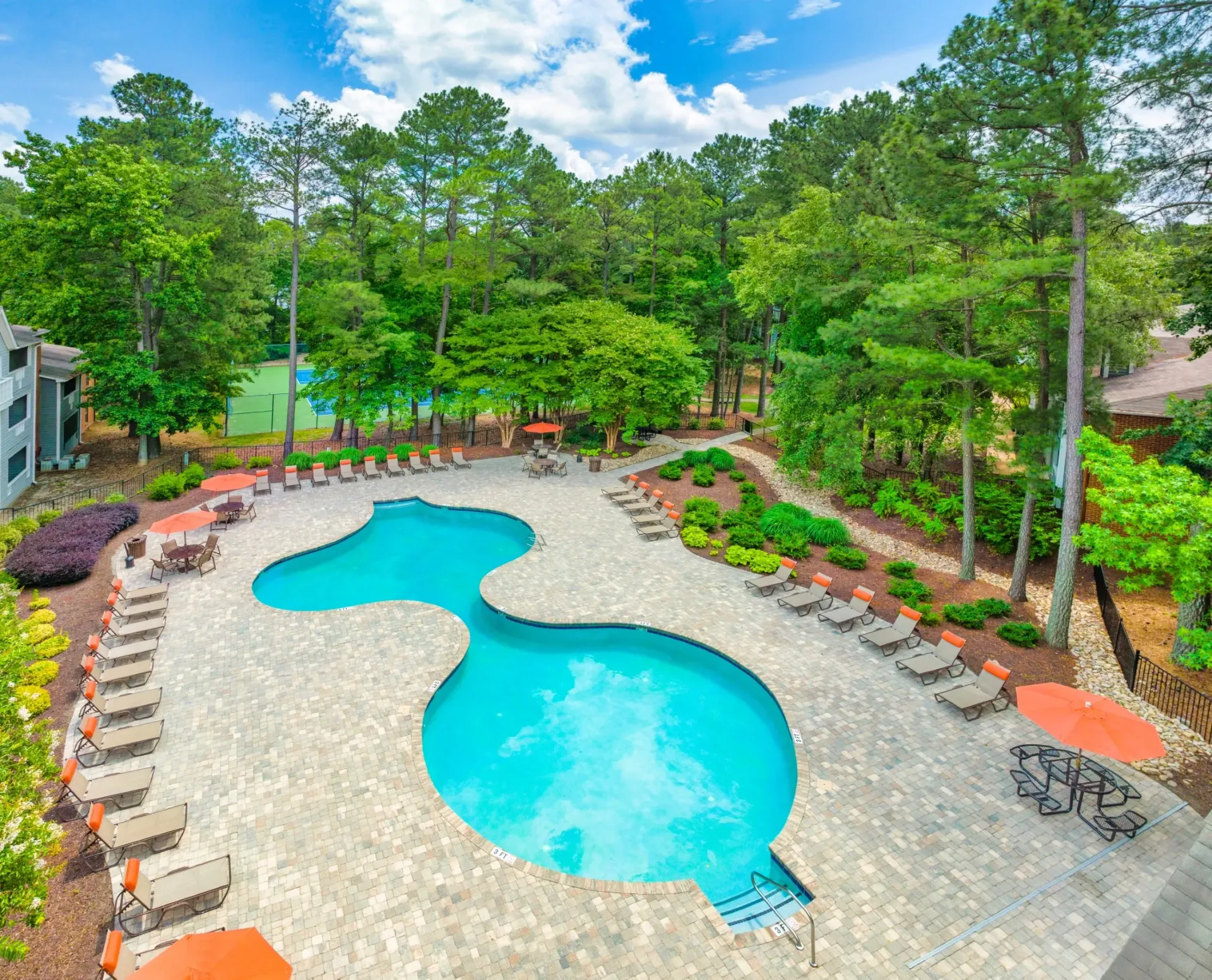 Outdoor swimming pool with lounge chairs and umbrellas, surrounded by trees and a tennis court.