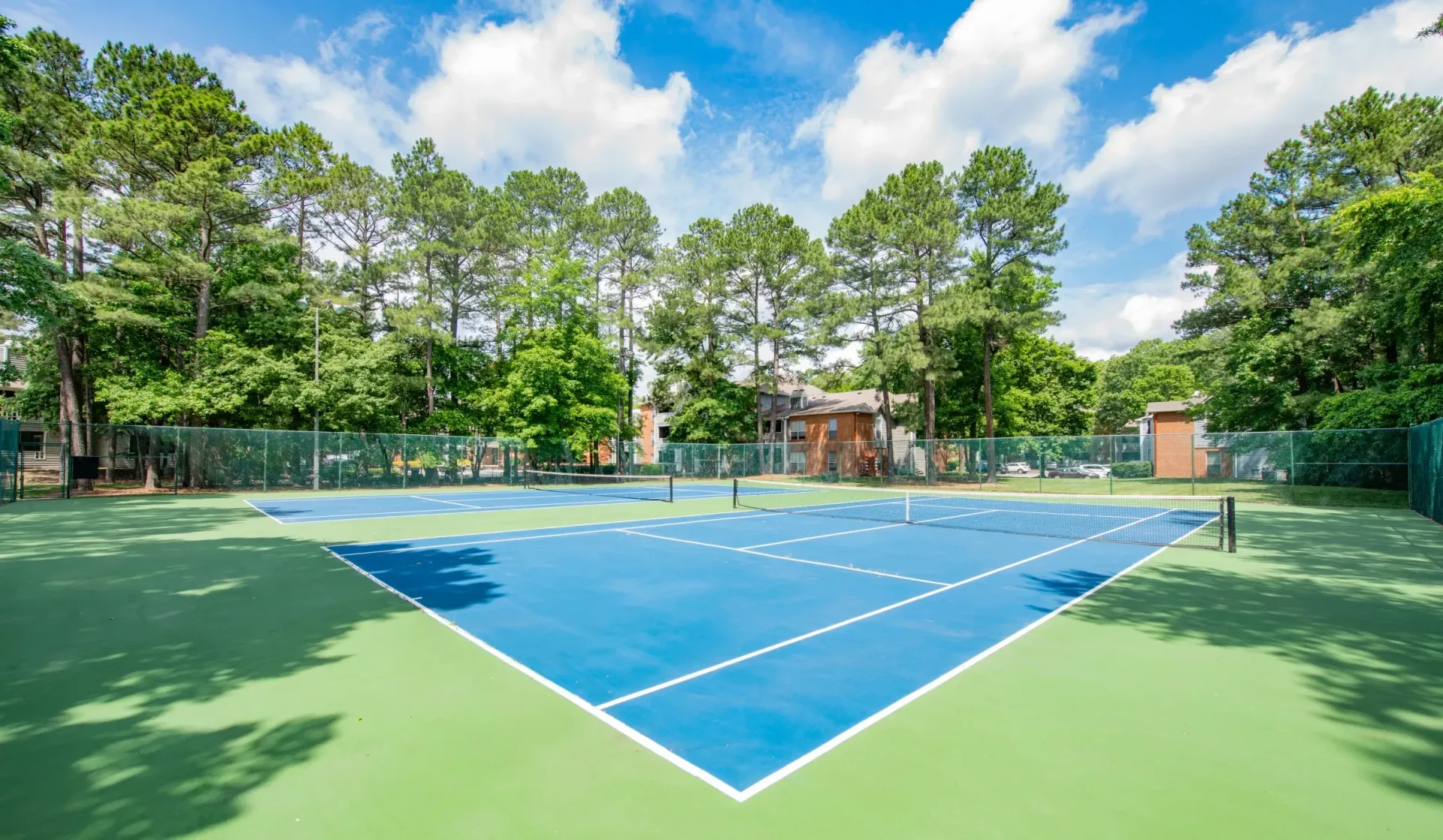 Two bright blue tennis courts with green surrounds and fencing, set against a backdrop of lush trees and a partly cloudy sky.