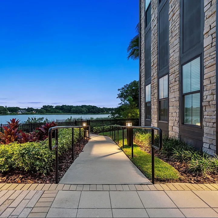 Charming lakeside apartment with a paved walkway, surrounded by lush greenery, overlooking a serene water view at dusk.