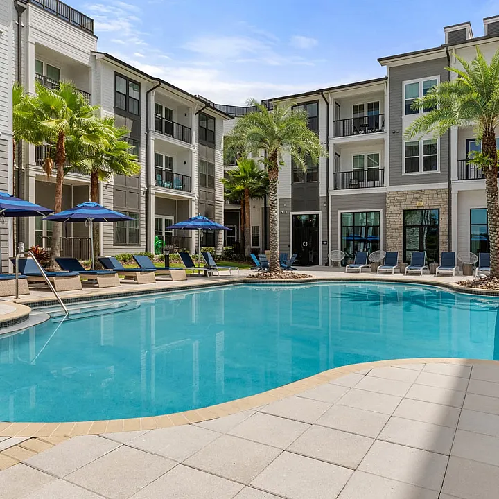 Luxurious apartment complex featuring a sparkling pool surrounded by modern architecture, palm trees, and relaxing lounge chairs under blue umbrellas.