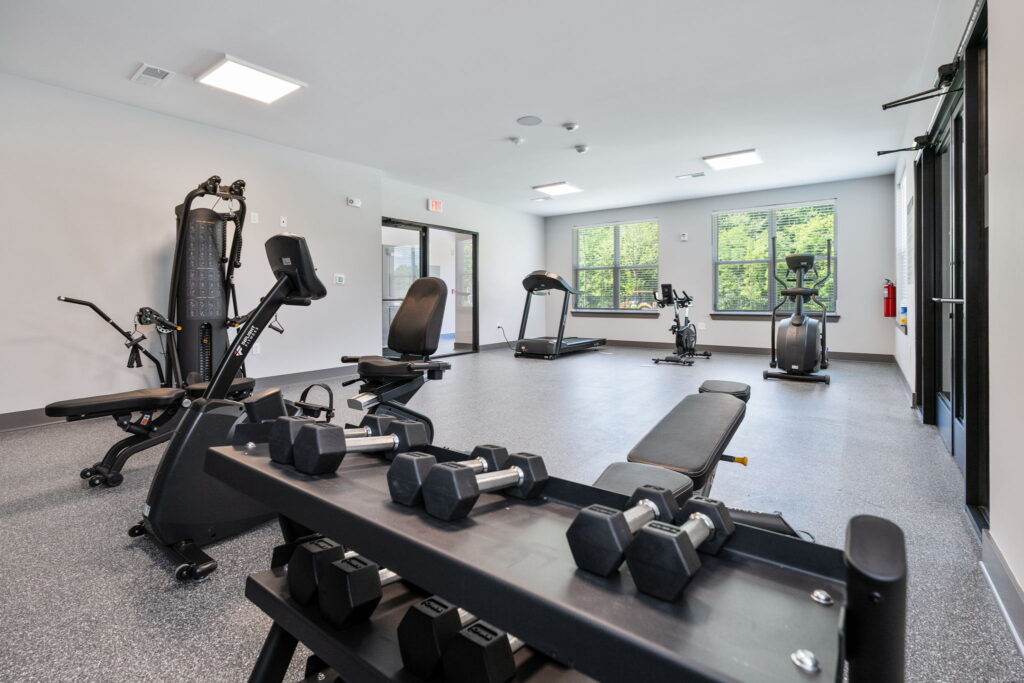 A modern gym with free weights, weight benches and cardio equipment. There are large windows that provide natural light and a view to the outside greenery.