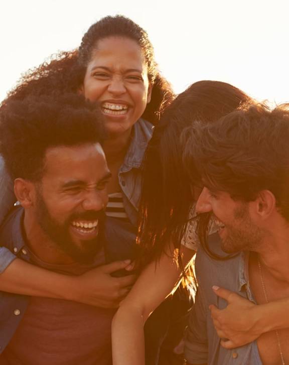 A group of friends are hugging and laughing together in a sunny outdoor setting.