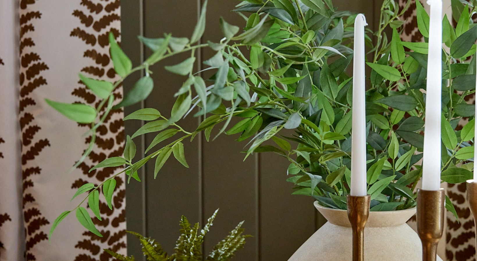 Decorative tabletop with greenery in a ceramic vase, brass candlesticks, and stacked books in a warm, modern interior setting