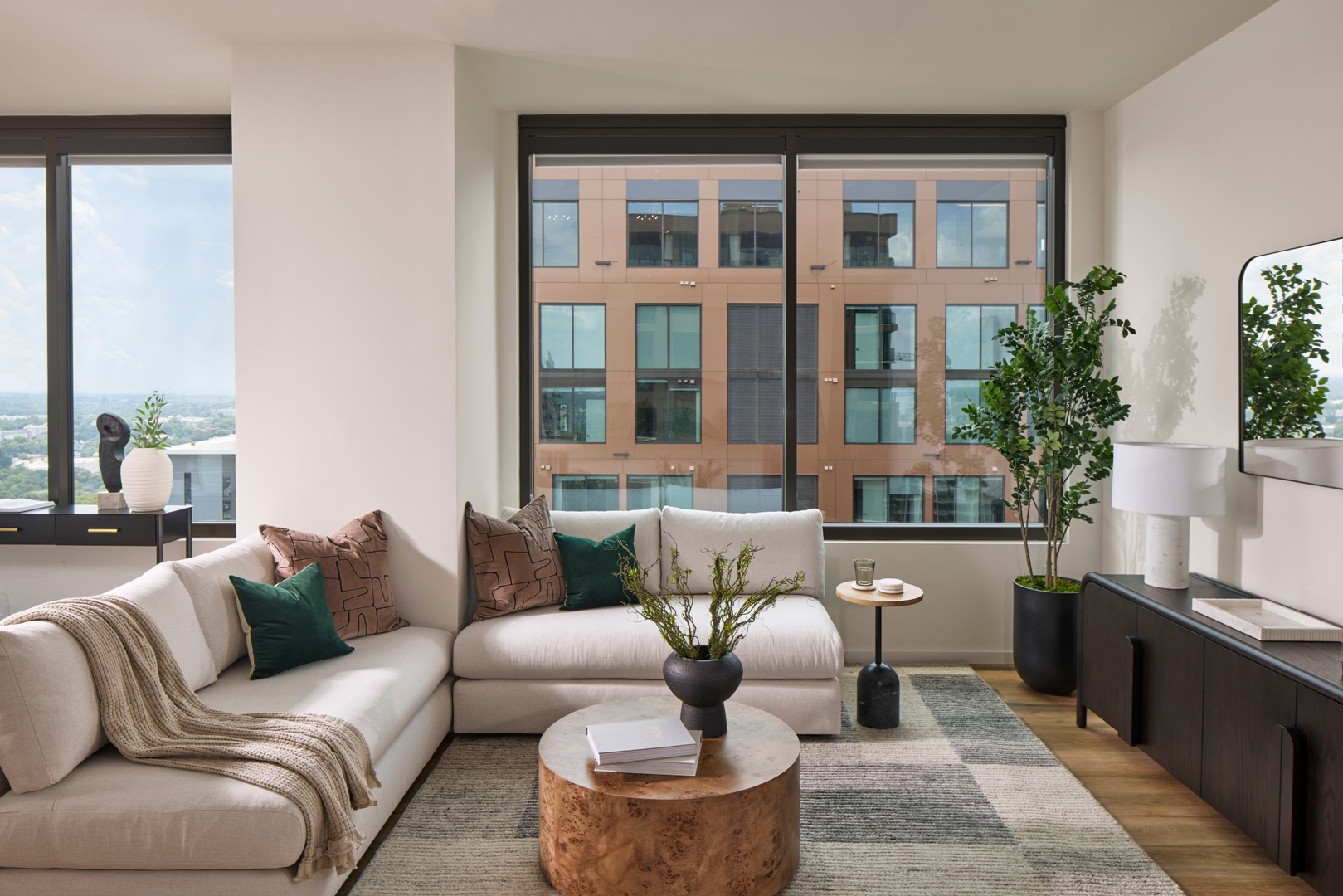 Bright living space featuring a cream sectional, wooden coffee table, decorative pillows, and large windows with city views.
