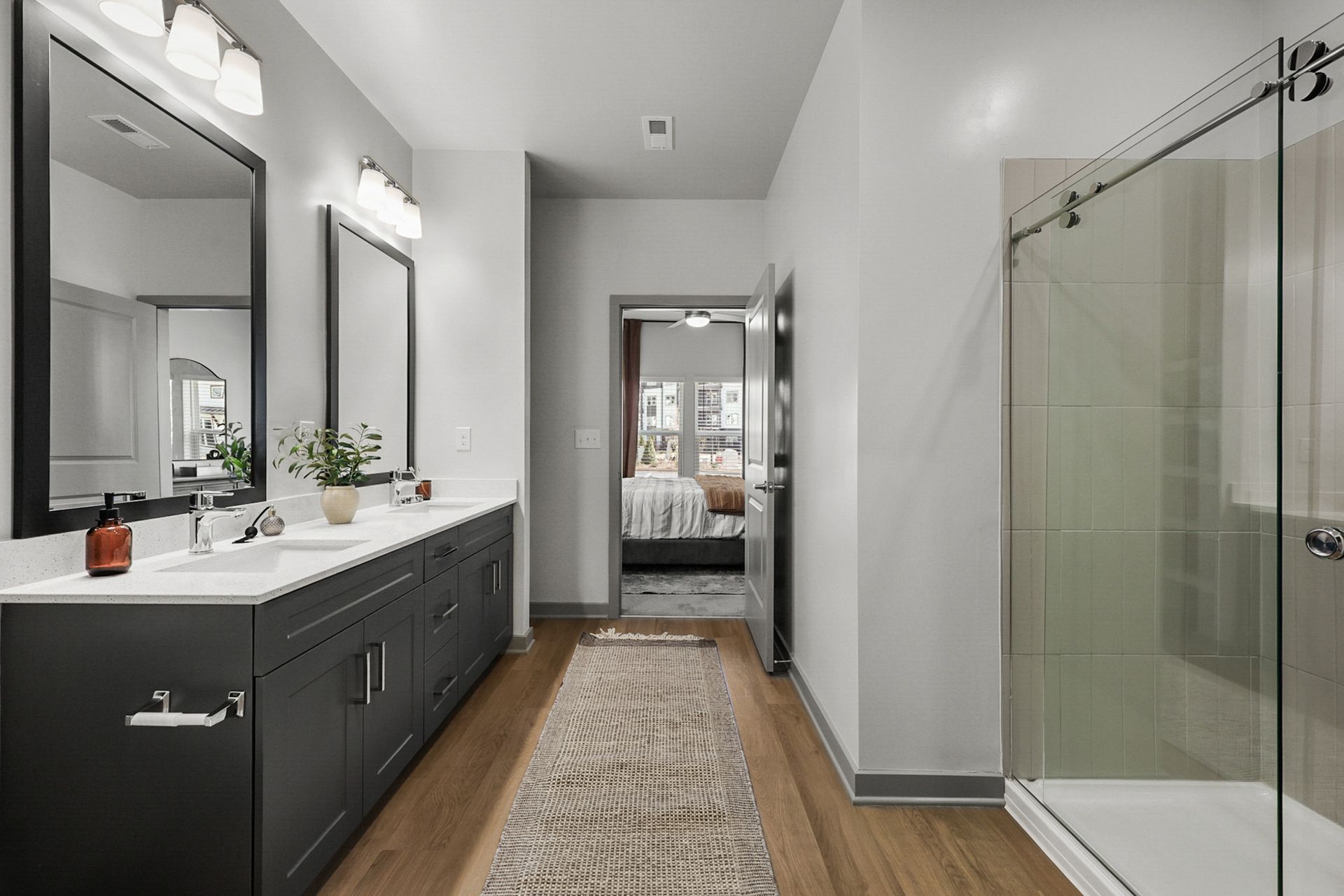 Modern bathroom with double vanity, large mirror, glass shower, and a runner rug leading to a bedroom.