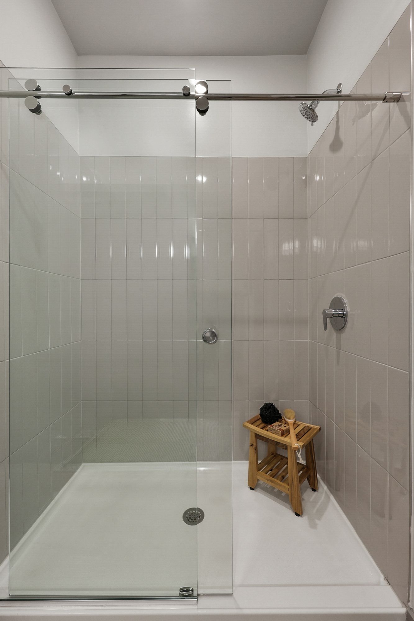 Glass shower stall with white tiled walls, sliding door, and a small stool holding toiletries.