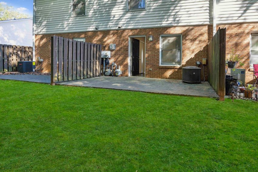 View of a backyard patio area with green grass, featuring a wooden fence on either side. The adjacent building has a brick exterior and several windows. Visible items include an air conditioning unit and some outdoor furniture. The scene is well-lit, showcasing a tidy outdoor space.