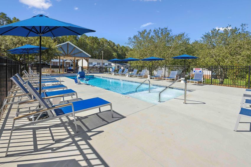 A swimming pool area featuring a clear blue pool with a shallow entrance, surrounded by lounge chairs and blue umbrellas. Green trees are visible in the background, and a building can be seen near the pool. The scene is bright and sunny, creating a relaxing outdoor atmosphere.