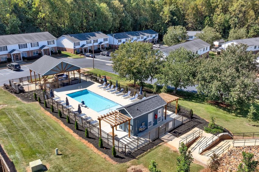 Aerial view of a residential community featuring a swimming pool surrounded by lounge chairs, a pergola, and landscaped green spaces. In the background, several apartment buildings can be seen, nestled among trees, depicting a serene and inviting atmosphere.