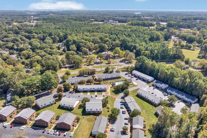 Aerial view of a residential area featuring multiple low-rise apartment buildings surrounded by green trees and lawns. The image shows a swimming pool area and nearby roads, with a dense forest in the background, indicating a suburban setting.