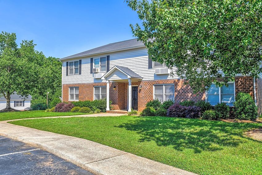 A well-maintained two-story brick and siding house with a welcoming front porch, surrounded by neatly trimmed lawns and colorful shrubs. The sky is clear blue, and the driveway curves gently in front of the house, emphasizing the property's inviting appearance.