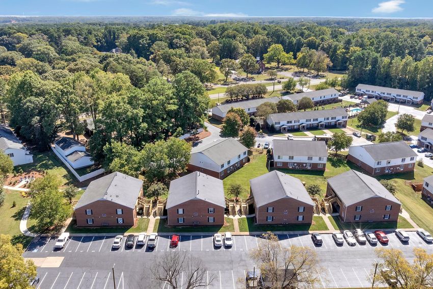 Aerial view of a residential neighborhood featuring several brick apartment buildings surrounded by green trees and lawns. There are paved parking areas in front of the buildings, and additional housing is visible in the background. The scene is set against a clear blue sky.