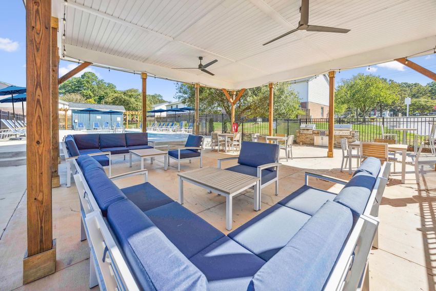 A covered outdoor seating area with blue couches and tables, overlooking a swimming pool surrounded by deck chairs. The space is well-lit and features ceiling fans, with greenery and a fence in the background.