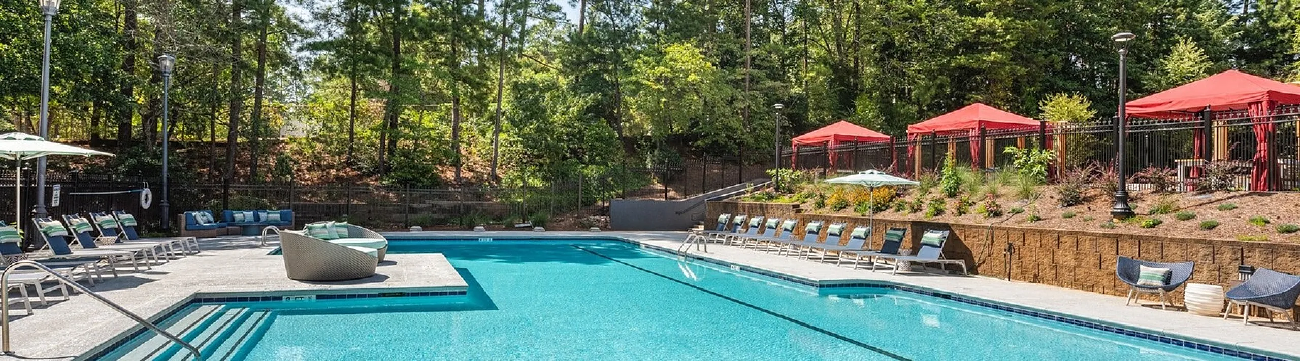 Inviting outdoor pool with sun loungers and vibrant red cabanas, surrounded by lush greenery and tall trees under a clear blue sky.