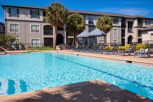 Resort-style swimming pool with lounge chairs and palm trees in front of apartment buildings.