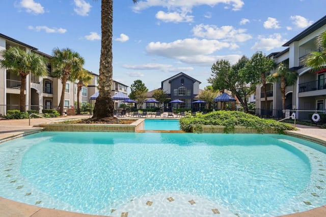 Resort-style swimming pool with lounge chairs and umbrellas, surrounded by palm trees and apartment buildings.