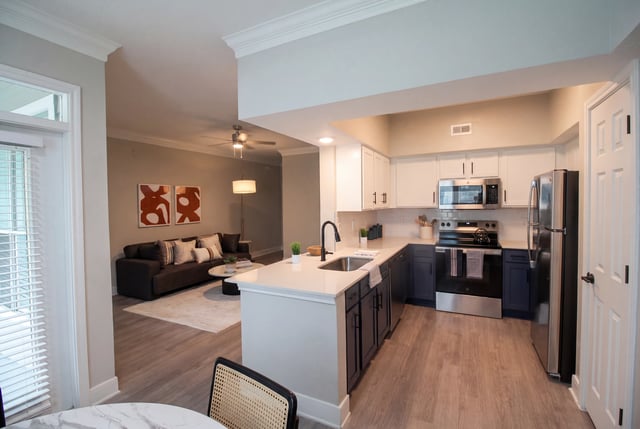 Modern kitchen and living room in apartment with dark blue cabinets, stainless steel appliances, and light wood floors.