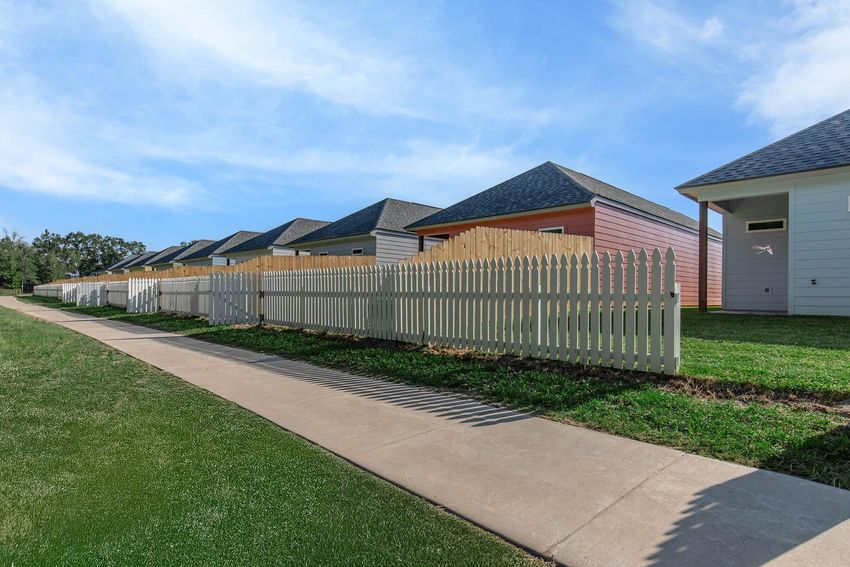 A row of colorful houses with wooden fences along a paved pathway. The houses feature gabled roofs and varying paint colors, set against a clear blue sky. Lush green grass borders the walkway, creating a pleasant suburban scene.