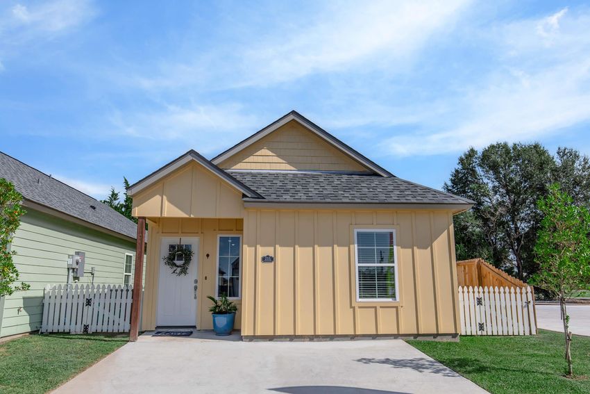 A charming yellow house with a gable roof, featuring a white front door and a wreath. The home has a small porch with a potted plant and is located in a neighborhood with green lawns and trees. A white picket fence is visible on one side, adding to the inviting curb appeal.