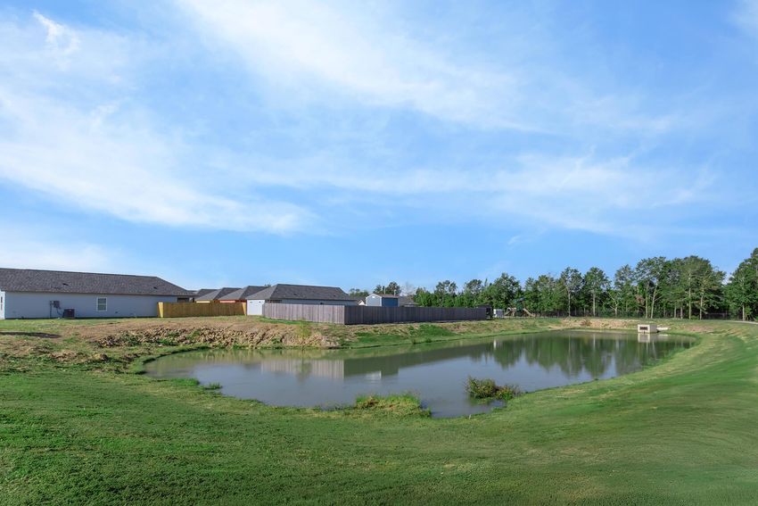 A serene landscape featuring a calm pond surrounded by grassy terrain. In the background, there are several houses with new development in progress. The sky is clear with a few wispy clouds, creating a peaceful suburban atmosphere.