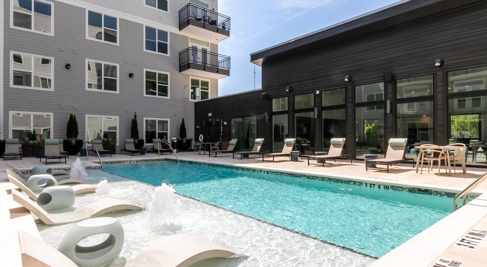 Luxury Apartments for Rent in Old Fourth Ward, GA - Tailor 4OW - Swimming Pool with In-Pool Loungers, In-Pool Stools, Sun Loungers, Dining Tables, Chairs, and View of the Clubhouse and Apartments.