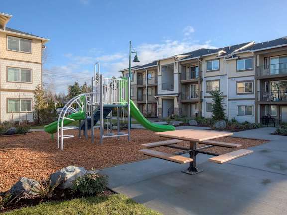 A playground with a green slide and a picnic table in front of apartment buildings.