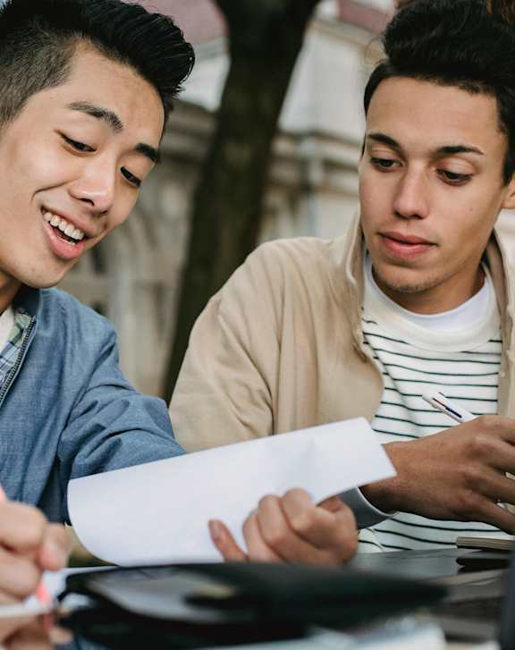Two men are sitting at a table, one is writing on a piece of paper while the other looks on.