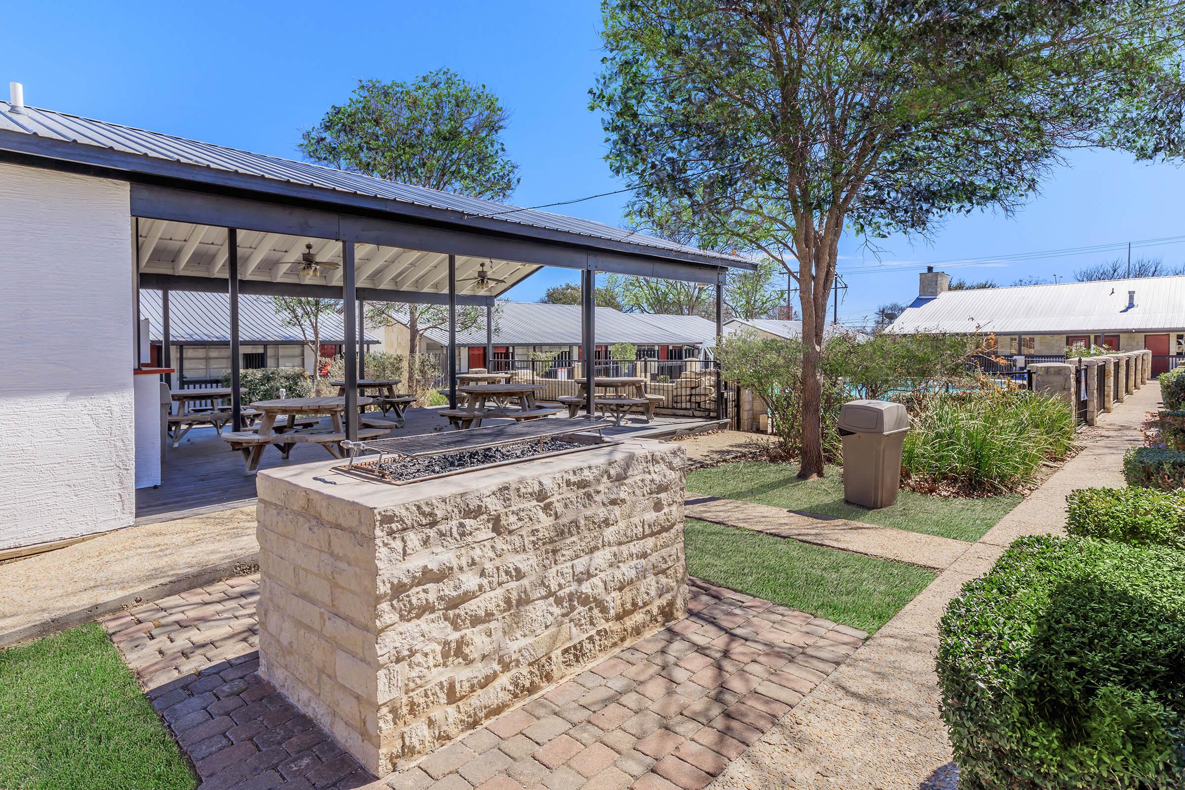 Outdoor seating area featuring picnic tables under a covered pavilion, a stone fire pit in the foreground, landscaped greenery, and a clear blue sky. The surrounding space includes a well-maintained path made of pavers and grass, with additional buildings visible in the background.