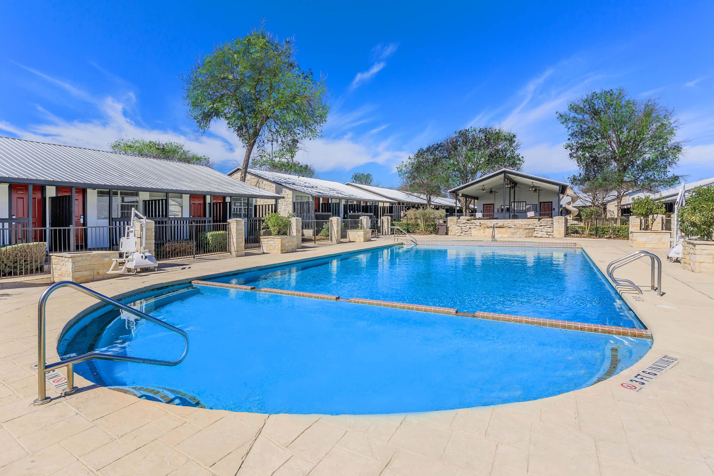 A clear blue swimming pool surrounded by a tiled deck, with lounge chairs and a small covered pavilion in the background. The area features several cabins with red doors and green trees, set against a bright blue sky with scattered clouds.